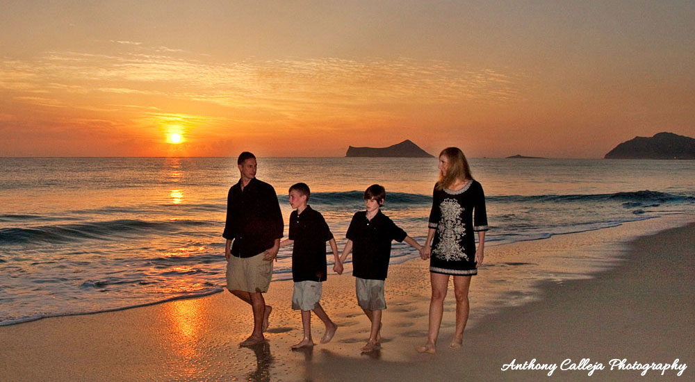 Family of four holding hands walking on the white sands of Waimanalo Beach at sunrise. Rabit Island and Makapuu lighthouse in the background